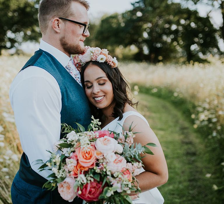 Groom in blue waistcoat and white floral tie with bride resting her head on the groom's chest with pink flower crown