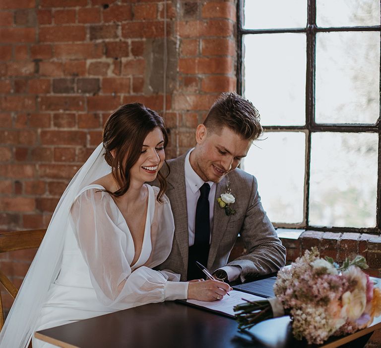 Bride wearing white fitted wedding dress with custom sheer puff sleeves signs the register with groom in puppytooth suit