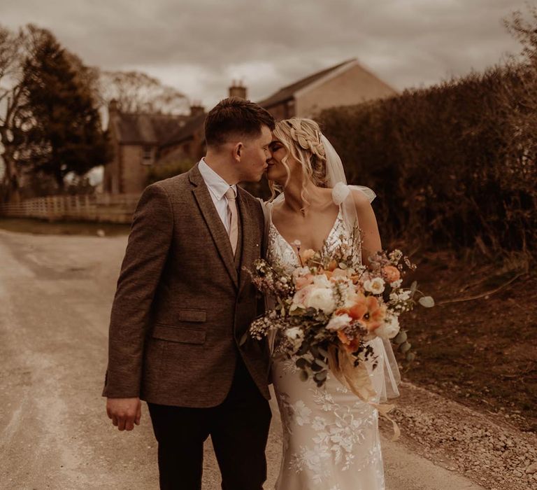 Groom in three piece brown suit kisses bride in lace Made With Love wedding dress as they walk around Eden Barn