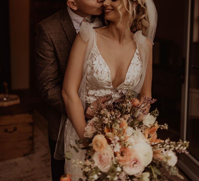 Groom in brown suit kisses the bride's cheek wearing Made With Love wedding dress and holding an autumnal wedding bouquet 