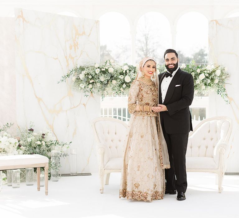 Bride & groom are surrounded by fresh white floral bouquets complete with green foliage in front of window 