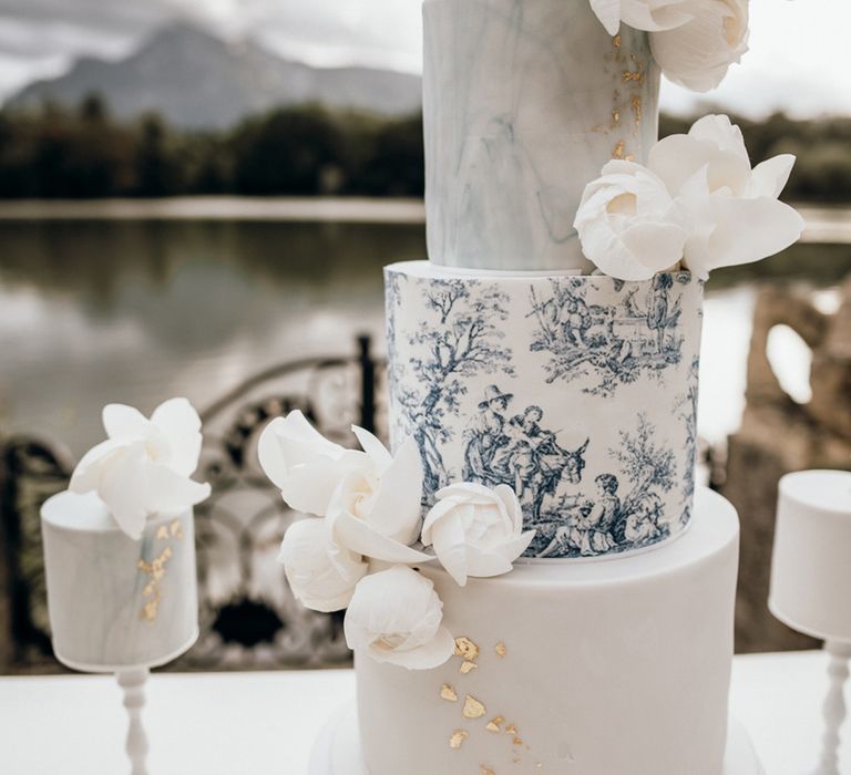 Three tier blue and white wedding cake with a marble layer, a patterned layer and a layer with gold flakes with white flowers