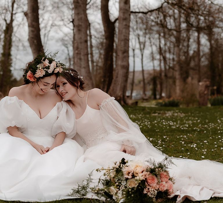 Bride in a floaty wedding dress with detachable sleeves sitting on the floor with her bride in a strapless wedding dress with puff sleeves and flower crown 