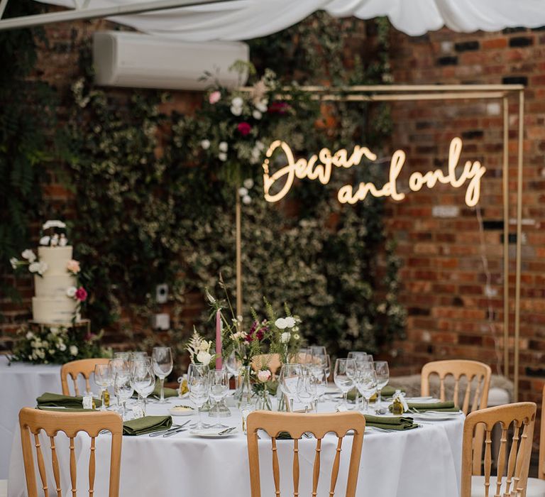 Personalised neon wedding sign hangs in front of floral foliage across wall and white table cloth lines round tables surrounded by wooden chairs