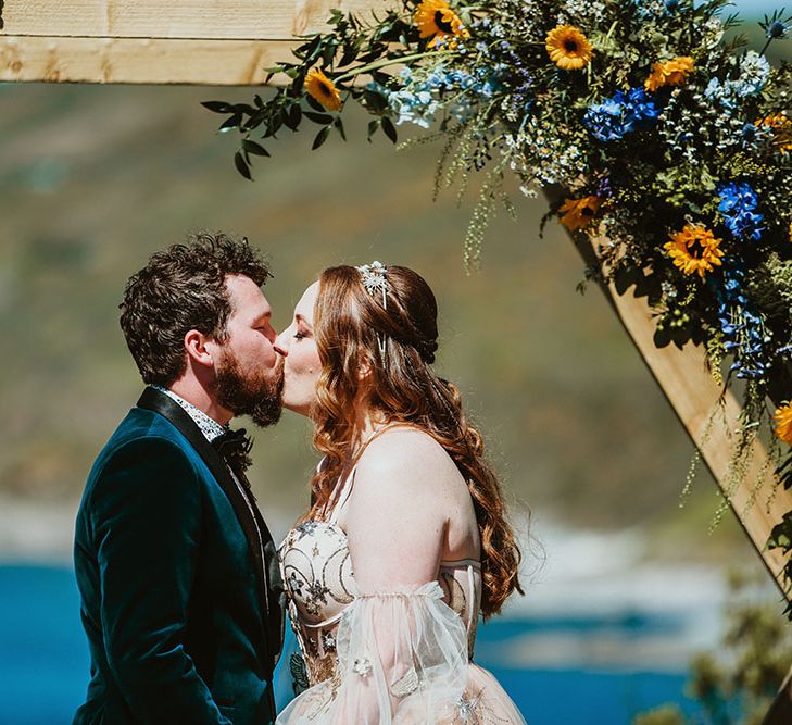 Bride & groom kiss beneath wooden arch finished with sunflower florals in front of the sea in Cornwall 