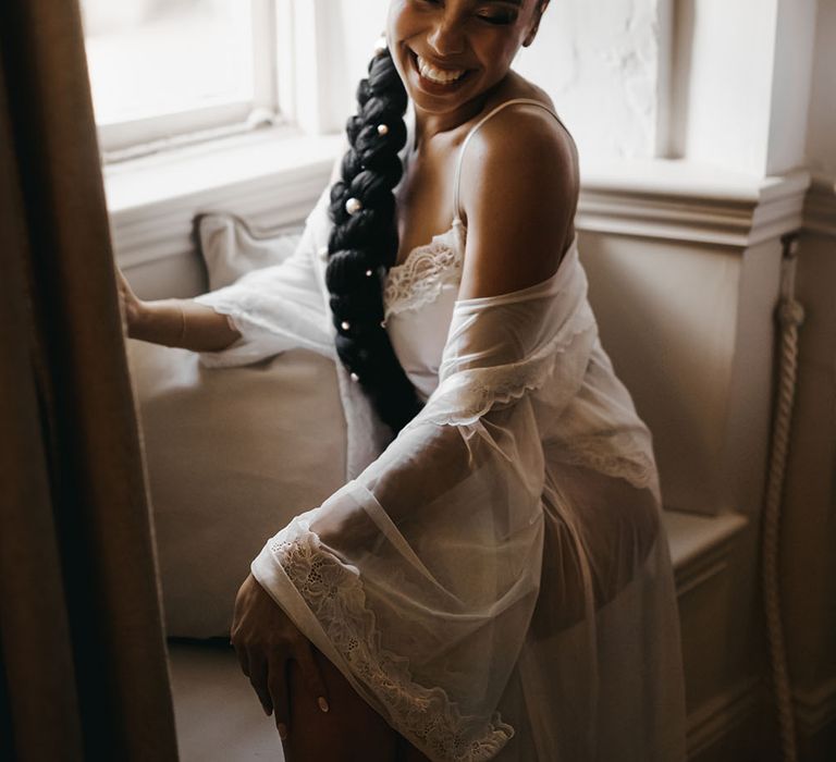 Black bride wears her hair braided with pearl hair accessories and silk lace white pyjamas on the morning of her wedding day