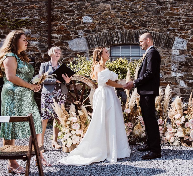 Bride and groom hold hands at the altar for their ceremony with a pink rose and pampas grass flower decoration 