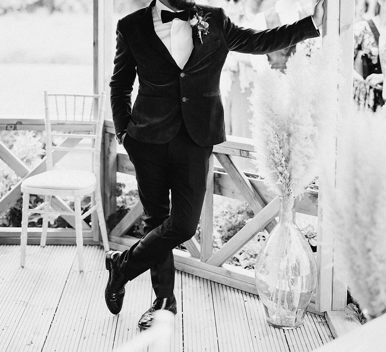 Groom in black tie leans stylishly against the gazebo waiting for the bride 