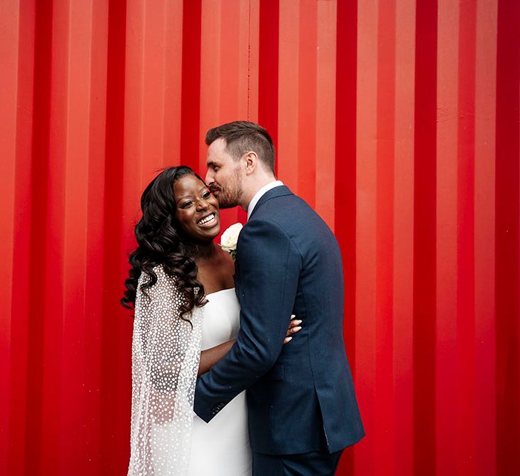 Groom kisses his brides cheek in front of bright red wall outdoors for Trinity Buoy Wharf wedding