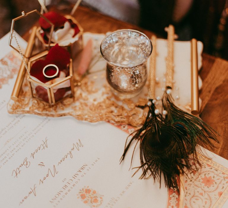 Gold wedding rings in ornate glass boxes beside colourful wedding stationery 