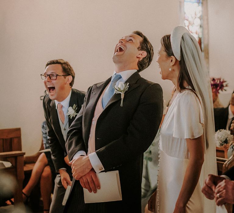 Groom in black morning suit with a pale blue tie and pink waistcoat laughing with the best man and bride at the altar 