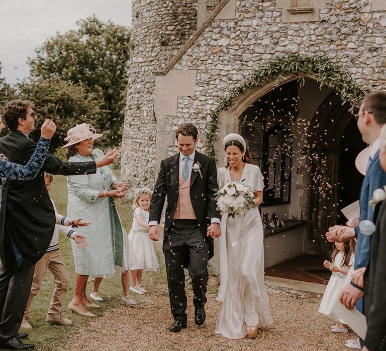 Groom in a traditional morning suit with a pink waistcoat and blue ties walking with the bride in a satin wedding dress and chunky headband exit the church to confetti