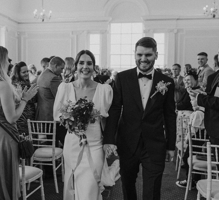 Bride in v neck wedding dress with puff sleeves and front slit and groom in black tux walking down the aisle together