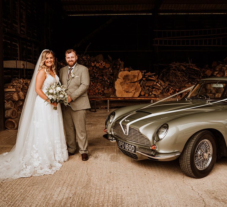 The bride and groom pose next to their Aston Martin grey wedding car transport 