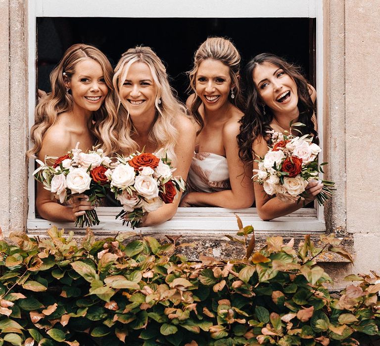 Bridesmaids holding rose bouquets lean out of the window together for a group photo