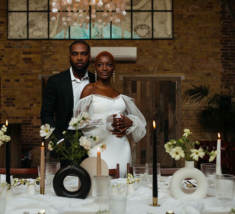 Bride in off shoulder wedding dress with lace puff long sleeves embracing groom in skinny black suit standing behind minimalistic wedding tablescape with black and white table decorations