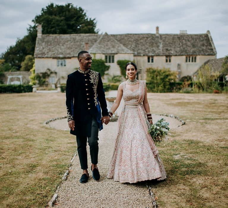 Panjabi Bride and Jamaican Groom walk hand in hand in front of Cotswolds wedding venue, Caswell House at multicultural wedding