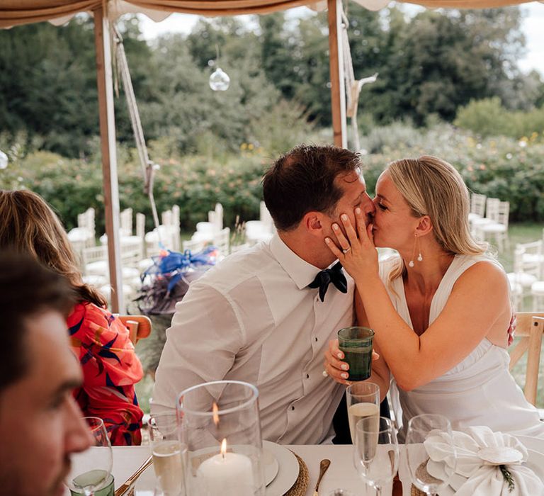 Groom in a white shirt sits with the bride as they share a kiss at their wedding reception in a marquee at Pythouse Kitchen Gardens 