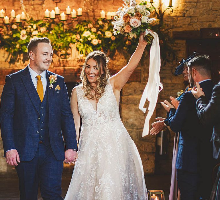 Bride in lace wedding dress holds up bridal bouquet while walking up the aisle with Groom in navy check wedding suit