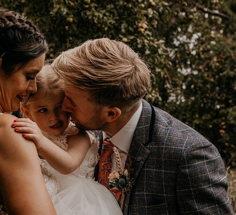 Groom in a checkered suit leans in to kiss the flower girl on the cheek in a white tulle dress being held by the bride with dark half up half down hair 