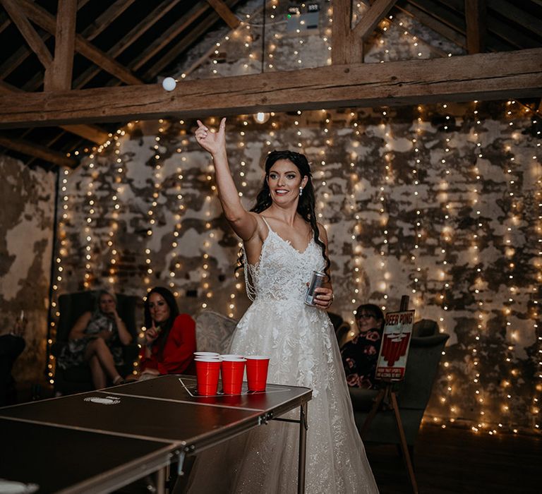 The bride in a lace wedding dress throws a white ball for beer pong game at wedding in a rustic barn with string fairy lights 