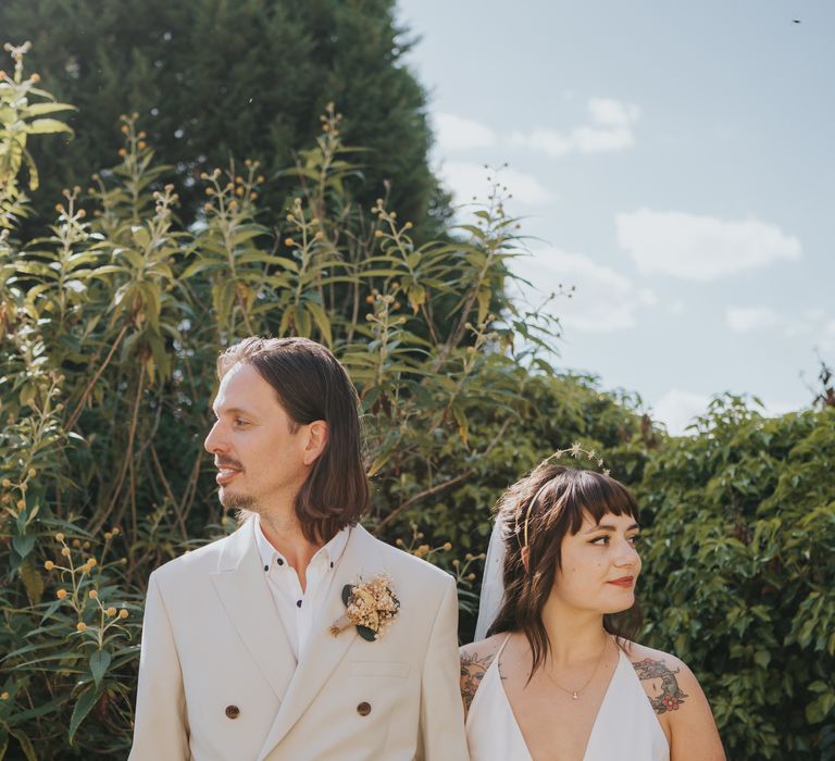 Groom in neutral coloured double-breasted ASOS suit and black high-top converse with dried flower and foliage boutonniere and bride in v-neck sleeveless ASOS wedding dress and gold halo headband with sparkly stars holding dried flower wedding bouquet with white bunny tails, green foliage, yellow dried gypsophila, lavender and baby’s-breath 