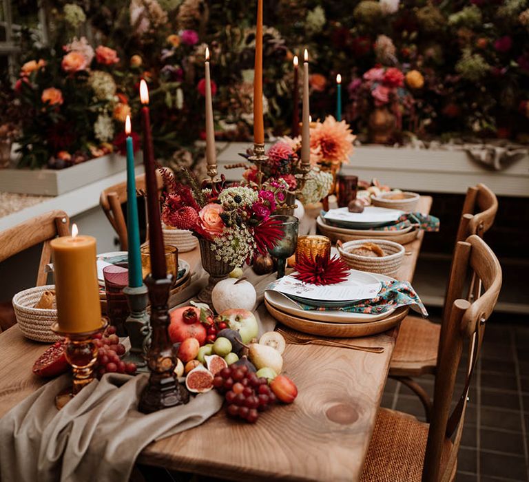 Neutral-toned tablerunner on rustic wooden table, orange, green and red coloured glasses, earthenware, seasonal fruit and vegetables, red, brown and green tapered candles in gold candlesticks and clear vases with autumnal coloured flowers