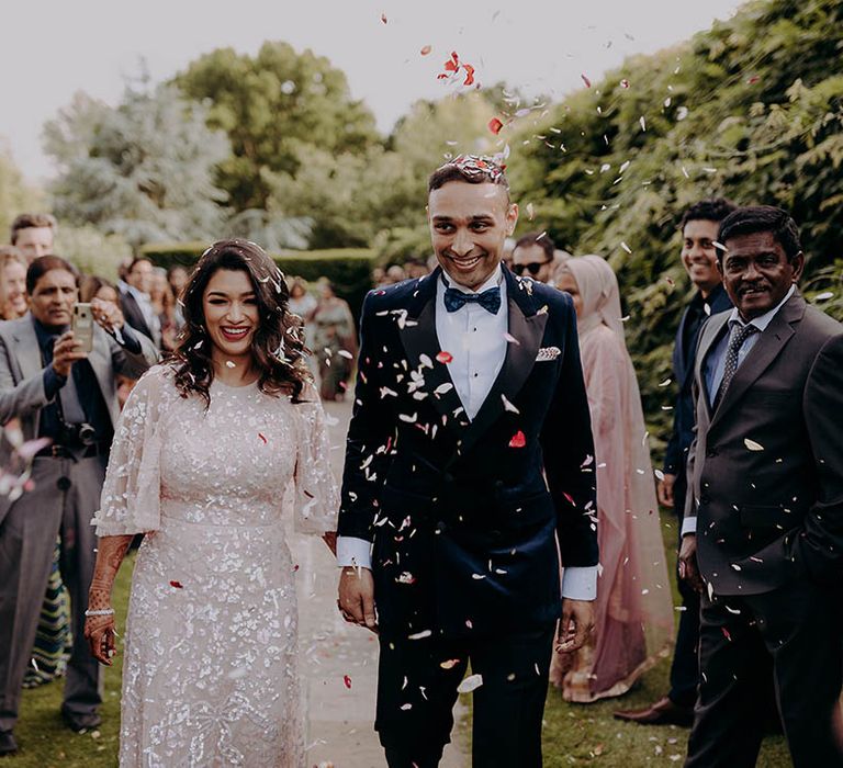 Groom in velvet tuxedo and bride in Needle & Thread dress walk through colourful confetti exit 