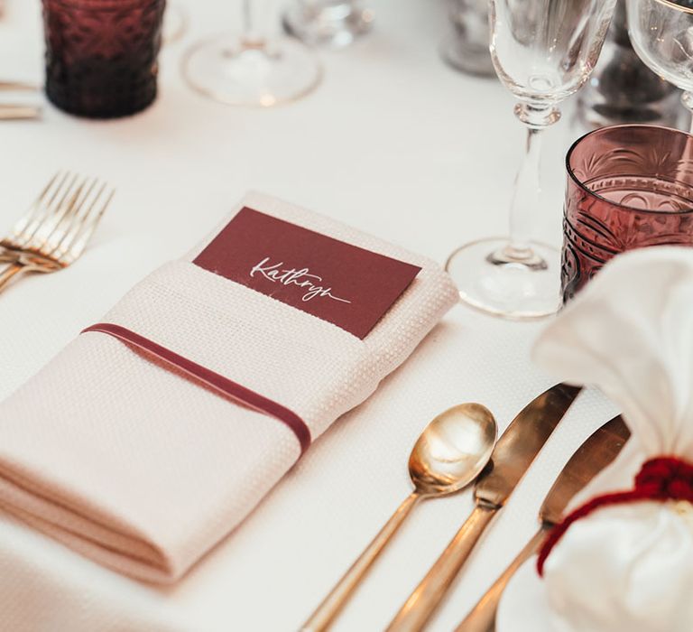 Gold cutlery with a burgundy place name, burgundy coloured glassware and ribbon with cream napkin for winter Christmas wedding  