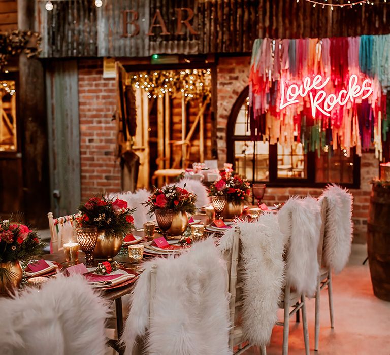 Rustic, boho luxe wedding reception room with tablescape with gold vases and hot pink plates, faux fur throws, streamer decoration ideas, hot pink neon wedding sign and fairy lights  