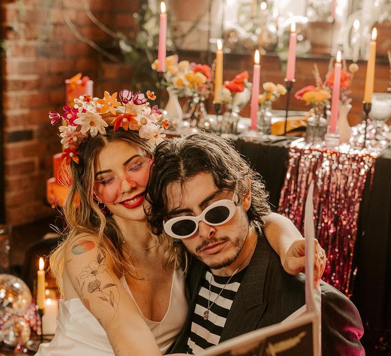 Groom in dark grey pinstripe blazer, large white sunglasses and black and white long sleeve t shirt reading tabloid style newspaper wedding stationery with bride in v neck satin wedding dress and pink bridal flower crown 