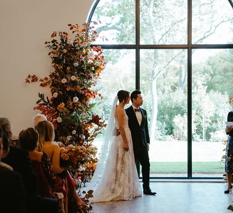 Bride & groom stand in front of large window beside burnt orange deconstructed arch flowers 