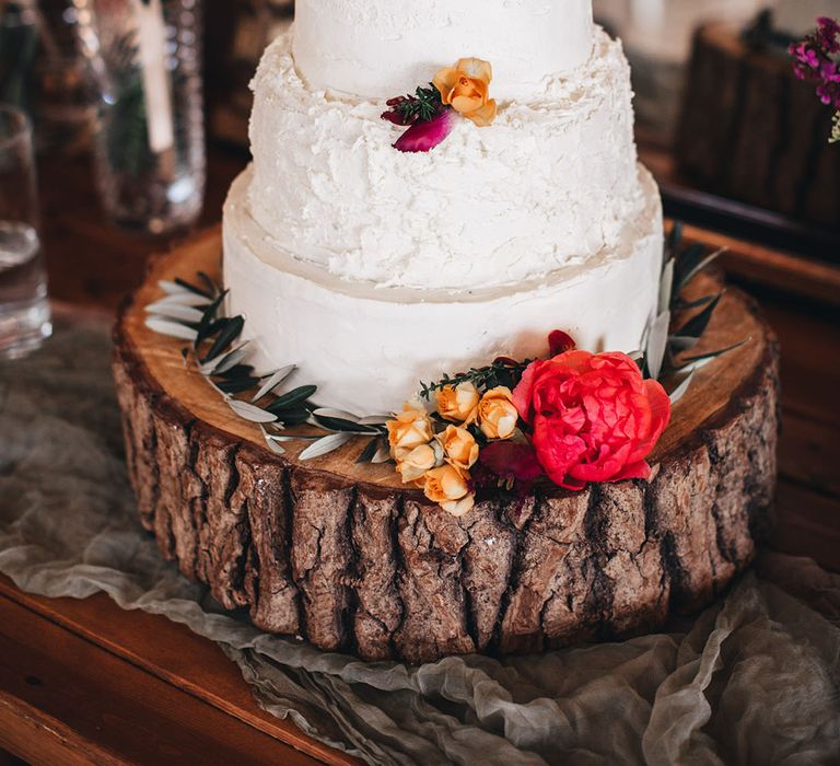 Three tier rustic wedding cake with white frosting on tree trunk cake stand with colourful flowers decorating the cake 