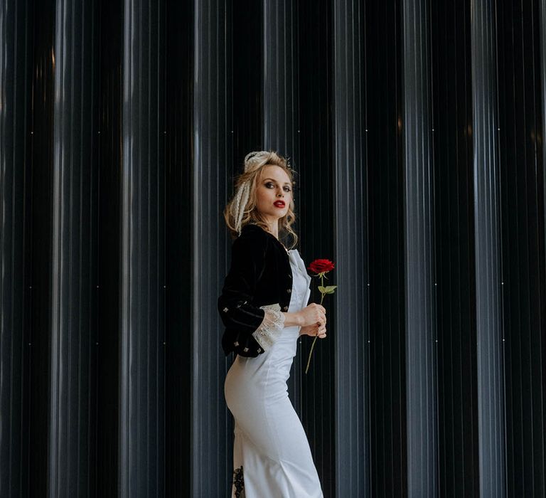 Bride in sculpted bandeau maxi wedding dress with black lace detailing on the train and black fitted jacket with cream lace sleeves holding single red rose 