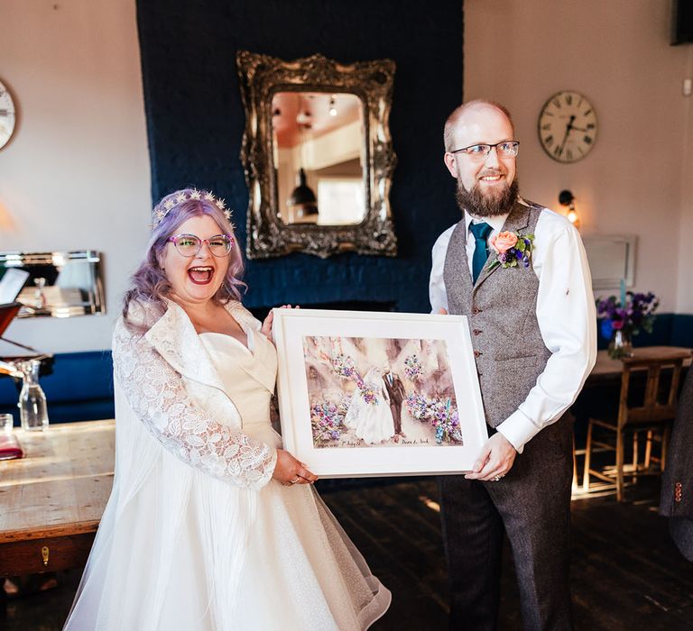 Bride in midi-length bridal gown with lace embroidered sleeves and polka dot wedding boots holding watercolour painting with groom in grey waistcoat, dark green tie and rose boutonniere 