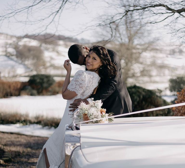 Bride in short sleeve lace wedding dress with groom in classic black tuxedo standing by a classic white wedding car in Yorkshire wedding venue