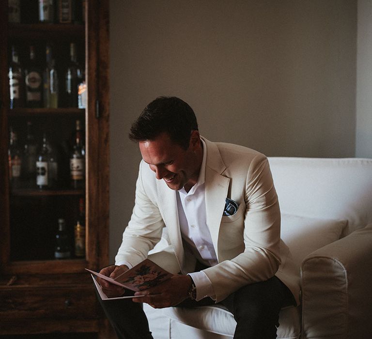 Groom in cream coloured suit reads card on the morning of his wedding day