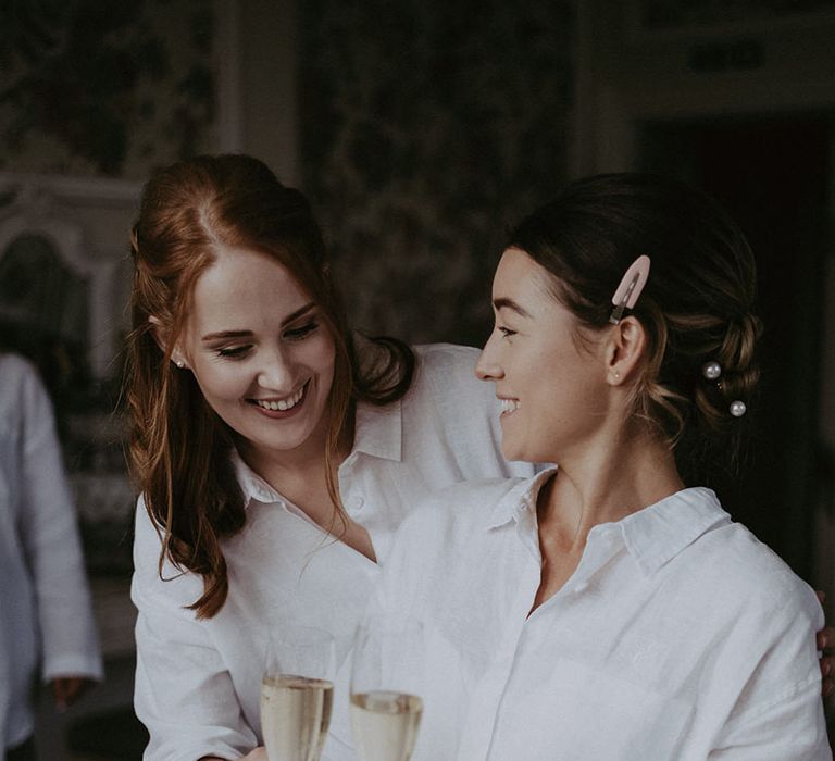 Bride wears white shirt and holds champagne beside bridesmaid on the morning of her wedding day