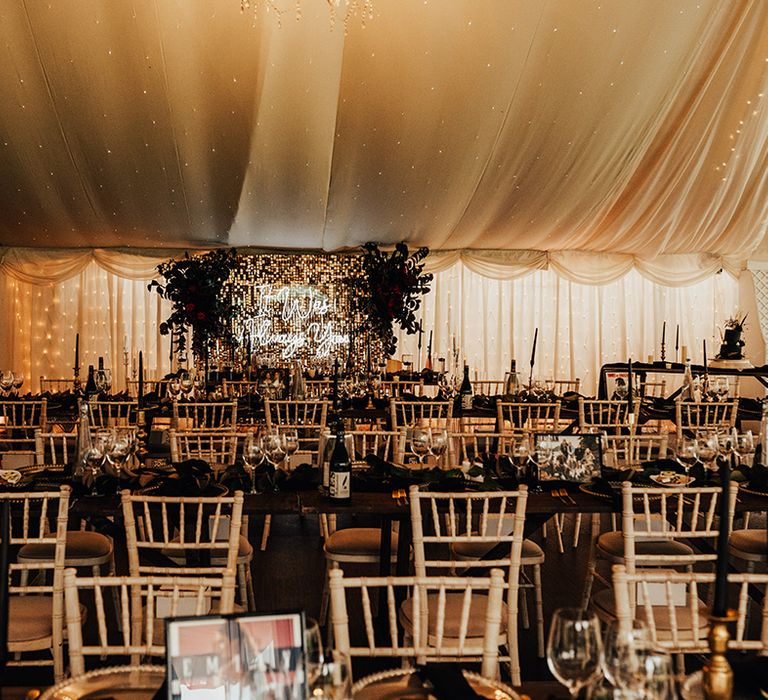 Marquee wedding with neon sign and a glitter wall with foliage and black accents and wedding decor including black taper candles, napkins, and place names