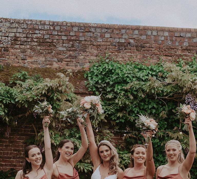 Bridesmaids in satin midi bridesmaid dresses in coral shade with the bride raising their bouquets into the air for bridal party photo 