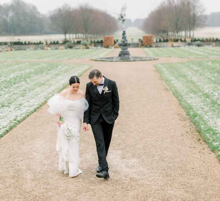 Groom in classic black tuxedo, black bowtie and boutonniere walking the grounds of St Giles House with bride in strapless wedding dress with puff tulle sleeves 