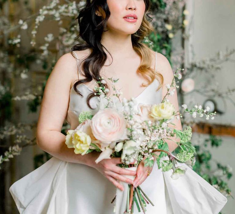 Bride in ivory satin strappy wedding dress holding rose, foliage and baby's-breath spring bouquet wearing large bridal headband 