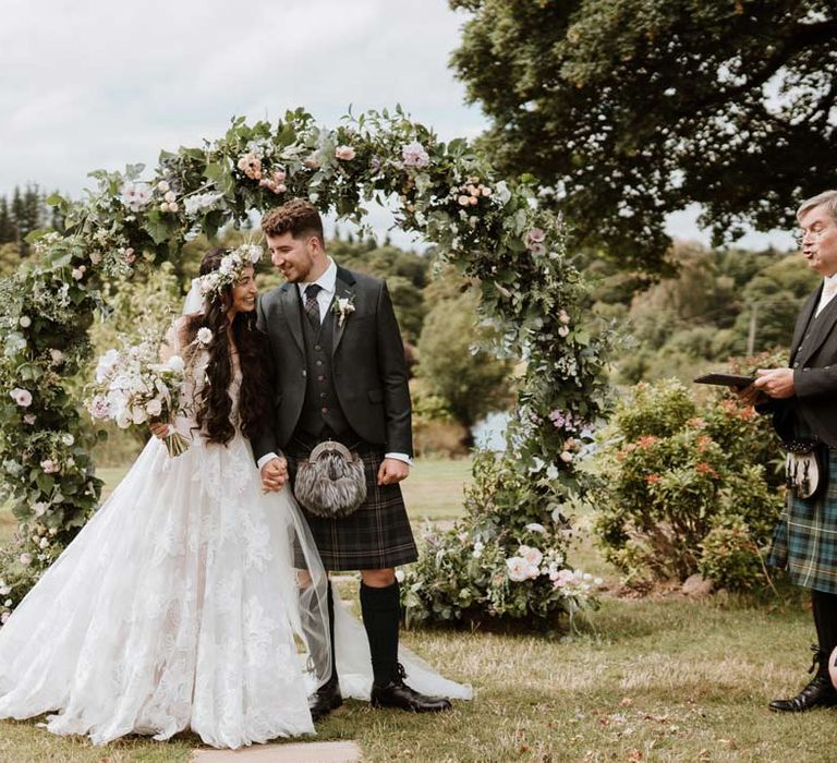 Bride wearing ethereal, celestial 3d appliqué sheer overlay wedding dress and bridal flower crown holding large white rose, peony, dried flower and foliage bridal bouquet standing with groom wearing dark grey blazer and waistcoat, dark tartan tie with white flower boutonniere, dark grey and blue tartan kilt and wedding sporran in front of circular flower arch 