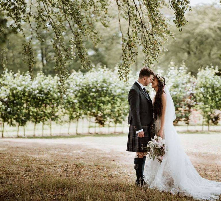 Groom in dark grey blazer and waistcoat, dark tartan tie and kilt with sporran embracing bride in 3d applique floral sheer overlay wedding dress with white garden rose flower crown 