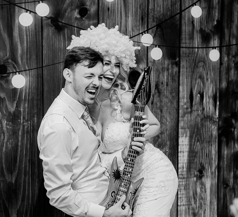 Groom posing with blow up guitar with the bride in a lace wedding dress wearing a fun wig for the wedding photo booth 