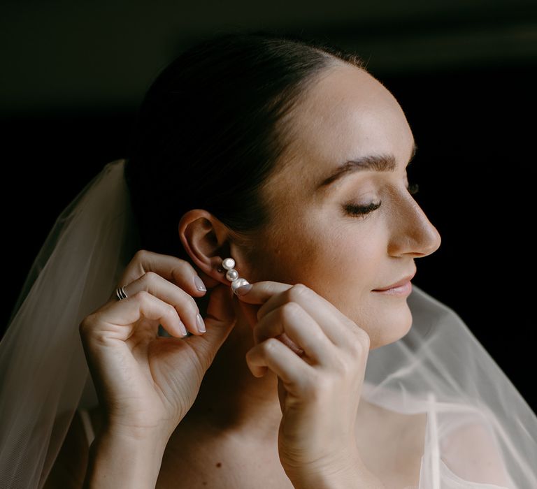 Bride in satin wedding dress puts in pearl earrings for her wedding day 