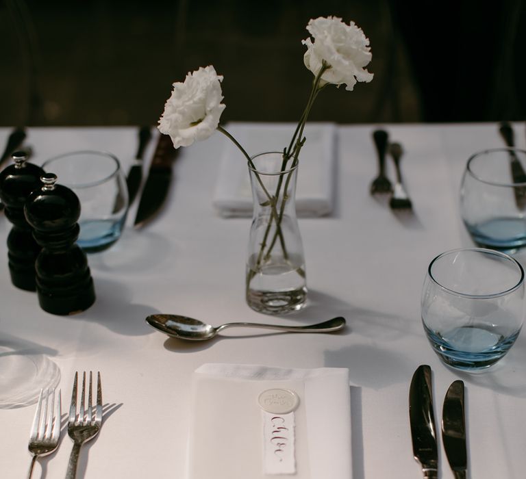 Minimal and modern wedding place setting with white wedding menu, napkins, and tablecloth with silver cutlery, blue ombre glassware and vase of white roses