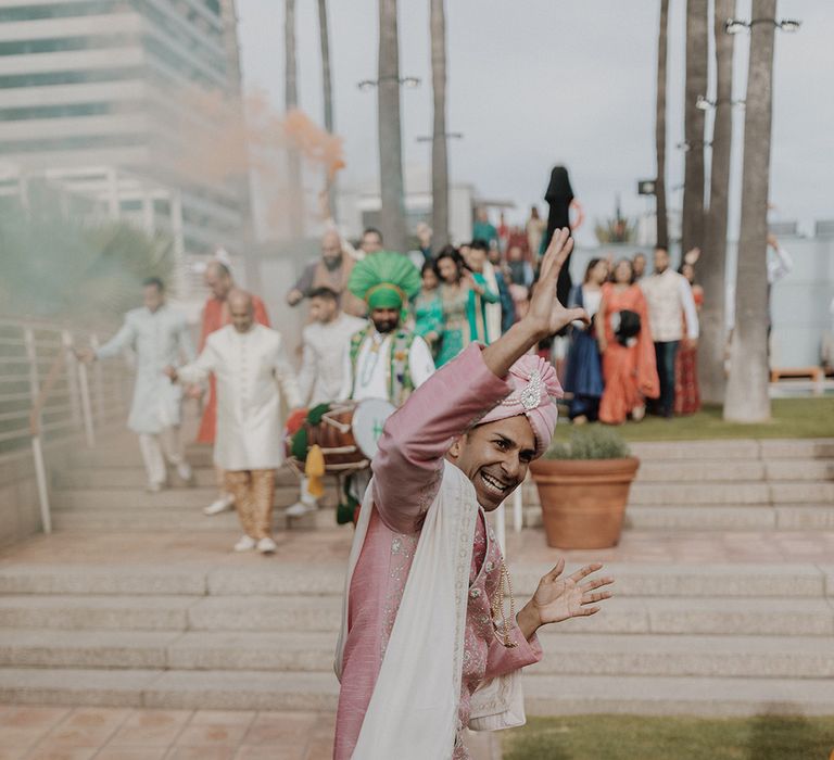 Groom in light pink sherwani, cream trousers and light pink groom shoes doing Indian dance with wedding guests doing colourful smoke bomb photoshoot in the background 