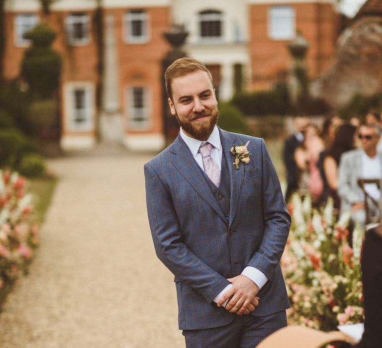 Groom in blue suit with pink paisley tie and pink floral buttonhole waiting for the bride at the end of the aisle 