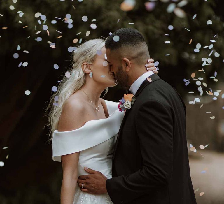 Bride in off the shoulder lace wedding dress holding colourful spring bridal bouquet kissing groom in classic three piece grooms tuxedo with colourful spring boutonniere at Frensham Hall doing colourful confetti photoshoot 